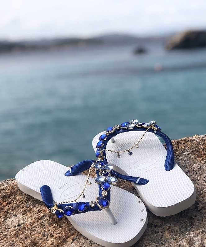 Sandalias blancas con detalles de piedras azules y doradas, sobre roca con mar de fondo.